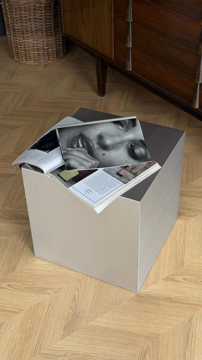 May include: A modern, square side table in a neutral beige color. The table has a minimalist design and is topped with open magazines featuring black and white photography. The table is placed on a wooden floor.