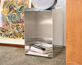 Könnte beinhalten: Ein silberner, würfelförmiger Beistelltisch mit einem grauen Lautsprecher oben. Der Tisch hat ein offenes Regal mit Büchern. Ein farbenfrohes gerahmtes Kunstwerk und ein Holzschrank befinden sich im Hintergrund.
