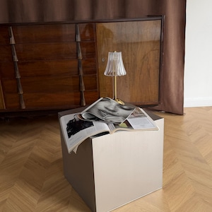 May include: A small, square, light gray side table with a gold-colored lamp and an open magazine on top. The lamp has a pleated glass shade. The background includes a wooden cabinet and a brown curtain.