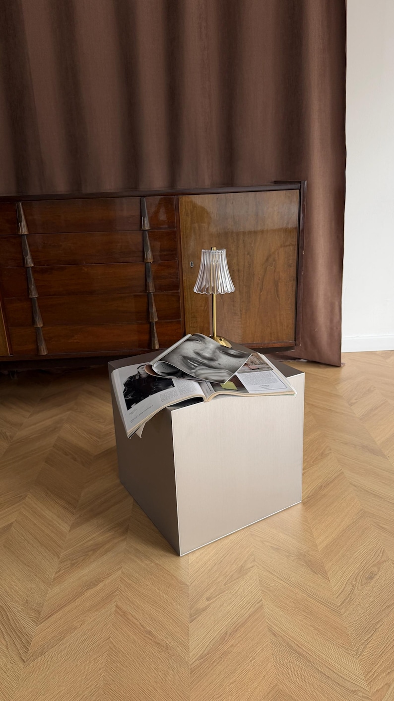 May include: A small, square, light gray side table with a gold-colored lamp and an open magazine on top. The lamp has a pleated glass shade. The background includes a wooden cabinet and a brown curtain.