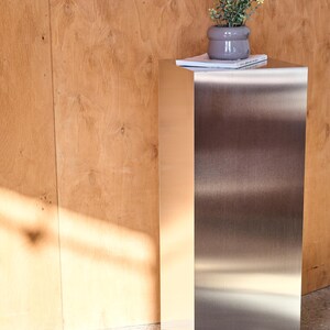 May include: A modern, stainless steel pedestal table with a small potted plant and a book on top. The table has a rectangular shape and a brushed metal finish. The background is a light brown wooden wall.