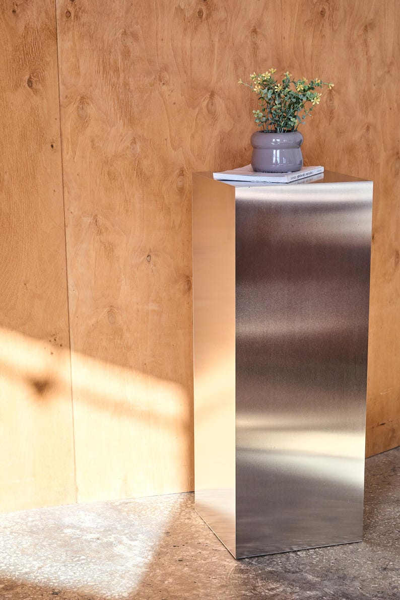 May include: A modern, stainless steel pedestal table with a small potted plant and a book on top. The table has a rectangular shape and a brushed metal finish. The background is a light brown wooden wall.