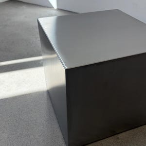May include: A modern, minimalist metal cube with a brushed silver top and dark gray sides. The cube is a solid, geometric shape, reflecting light and shadow. The design is simple and contemporary, suitable for various interior styles.