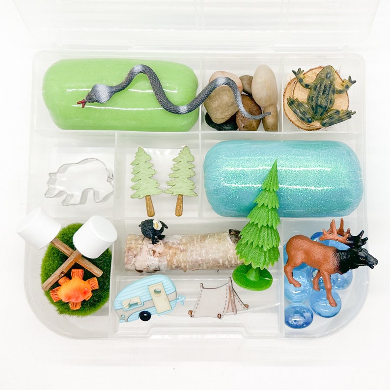 May include: A clear plastic storage box with nine compartments filled with various camping themed toys. The compartments contain green play dough with a snake, a brown bear, two green trees, a blue play dough with a green tree, a log with a black bird, a campfire with marshmallows, a blue camper, a green tent, and a brown elk with blue stones.