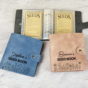 以下が含まれることがあります： 「Sophia's SEED BOOK」と「Bianca's SEED BOOK」の文字が入った青とピンクのシードブック。各本には花の模様とボタン留めが付いています。背景には、灰色のシードオーガナイザーが開いています。