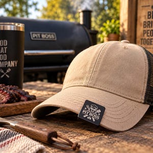 May include: A tan baseball cap with a black mesh back and a black rectangular patch on the brim featuring the Pit Boss logo, which includes a chef-hatted pig, crossed grilling utensils, and the text Pit Boss with a small flame icon below.