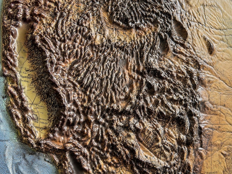 May include: A detailed relief map of a mountainous region, likely the western United States. The map features textured, raised areas in shades of brown and tan, with visible geographical features. The map has a vintage aesthetic, with some text visible.
