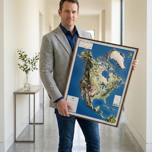 May include: A framed 3D relief map of North America, showcasing the continent's topography with detailed textures and colors. The map is set against a blue background and framed in a dark wood. The map is held by a person.