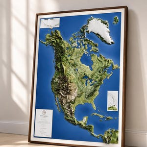 May include: A framed 3D relief map of North America, showcasing the continent's topography with detailed green and brown landmasses against a blue ocean background. The map is encased in a dark wooden frame with a white border, leaning against a wall.