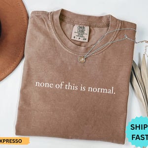 May include: A brown t-shirt with the phrase "none of this is normal" printed in white. The shirt has a crew neck and a small tag on the upper left. A silver necklace with a pendant is draped on the shirt.