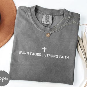 May include: A gray t-shirt with the text "WORN PAGES, STRONG FAITH" and a cross graphic. A gold necklace with a pendant is on the shirt. A brown hat and dried leaves are in the background. The shirt is labeled "Pepper".