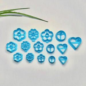 以下が含まれることがあります： さまざまな花とハートの形をした、水色のプラスチック製クッキーカッターのコレクション。カッターは白い表面に配置され、左上隅にはいくつかの緑の葉があります。形状には、花、円、ハートが含まれます。