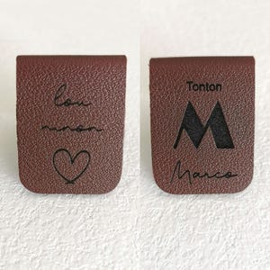 May include: Two brown rectangular leather labels with rounded bottom corners. The left label features the cursive text "lou" and "manon" above a heart outline. The right label displays the text "Tonton", a large letter "M", and "Marco".