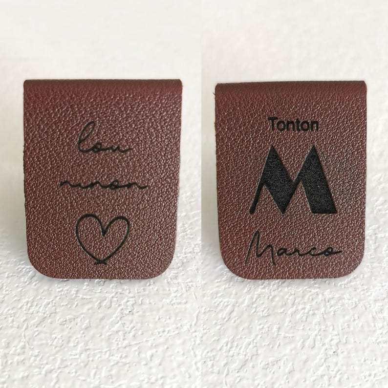May include: Two brown rectangular leather labels with rounded bottom corners. The left label features the cursive text "lou" and "manon" above a heart outline. The right label displays the text "Tonton", a large letter "M", and "Marco".