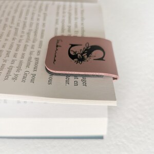 May include: A dusty rose magnetic bookmark clipped onto the top of a book page. The bookmark features a black printed design of the letter S decorated with floral elements and the name Sabrina written in a cursive script along the left side.