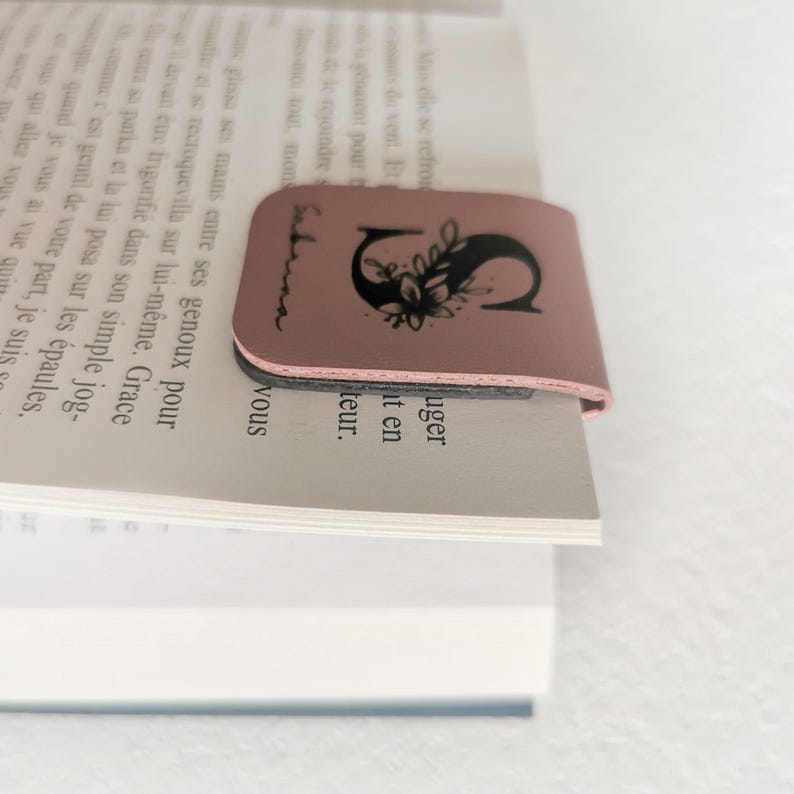 May include: A dusty rose magnetic bookmark clipped onto the top of a book page. The bookmark features a black printed design of the letter S decorated with floral elements and the name Sabrina written in a cursive script along the left side.