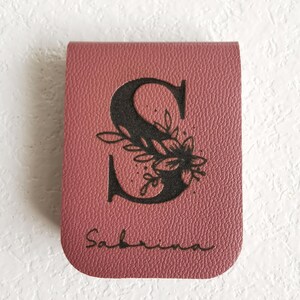 Marque-page en cuir bordeaux avec la lettre "S" ornée d'un motif floral noir. Le nom "Sabrina" est écrit en cursive en bas. Le marque-page est rectangulaire avec des coins arrondis.