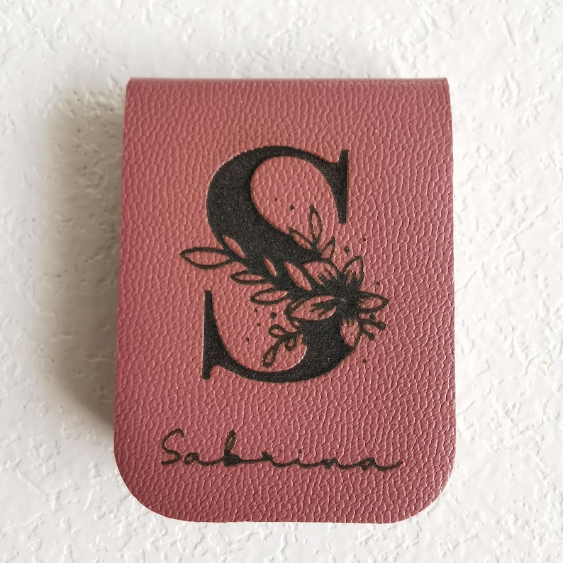 Marque-page en cuir bordeaux avec la lettre "S" ornée d'un motif floral noir. Le nom "Sabrina" est écrit en cursive en bas. Le marque-page est rectangulaire avec des coins arrondis.