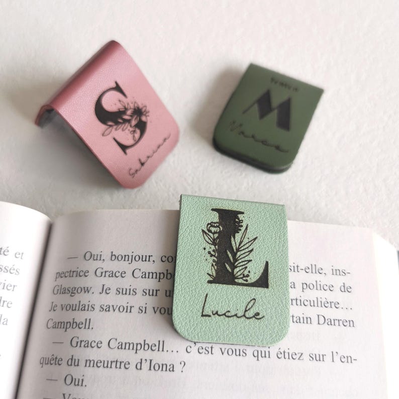 May include: Three magnetic bookmarks in pink, olive green, and light green. The pink one features a floral letter S and the name Sabrina. The olive green one shows a letter M and the name Marco. The light green one displays a floral letter L and the name Lucile.