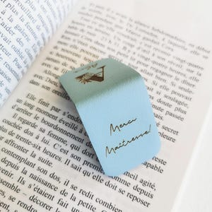 May include: A light blue magnetic bookmark with a textured surface, featuring a gold-colored graphic of a flower and the French text "Merci Ma&icirc;tresse!" printed in a cursive script, resting on the open pages of a printed book.