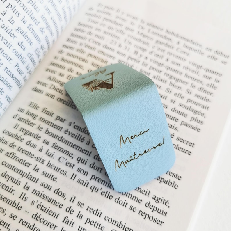 May include: A light blue magnetic bookmark with a textured surface, featuring a gold-colored graphic of a flower and the French text "Merci Ma&icirc;tresse!" printed in a cursive script, resting on the open pages of a printed book.