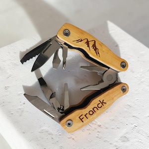 Op de afbeelding: Een multitool met een licht houten handvat en diverse roestvrijstalen gereedschappen. Het handvat is gegraveerd met een bergontwerp en de naam "Franck". De gereedschappen omvatten een zaag, mes, schroevendraaier en tang. De multitool is open.