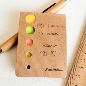 Può includere: Un blocco note Post-it marrone con note adesive colorate visibili attraverso ritagli circolari. Il blocco note ha testo in francese, tra cui "POST-IT pour ne rien oublier... même les PRÉNOMS!" Un righello e una penna di legno sono anche nell'immagine.
