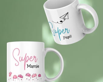 May include: Two white ceramic mugs. One mug has the words "Super Papi!" in blue script with a paper airplane illustration. The other mug has "Super Mamie" in pink script with pink flower illustrations.