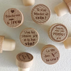 Pode incluir: Uma coleção de rolhas de garrafa redondas de madeira com bases de cortiça de cor clara. Cada topo de madeira apresenta texto em francês gravado a laser e pequenos ícones, incluindo corações, taças de vinho e saca-rolhas. Os designs incluem frases como Bouche-moi le trou, Tu m'en bouches un coin, Vive les copains et le bon vin, Bouche mais pas PERDU, La tête dans le cru e Amour en bouteille.