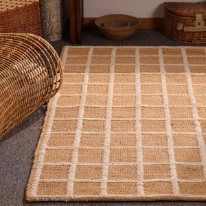 May include: A rectangular rug with a grid pattern. The rug is made of natural fibers, with a light brown color and off-white grid lines. The rug is placed on a gray surface. The text "Artisan Vibe Rugs" is at the bottom.