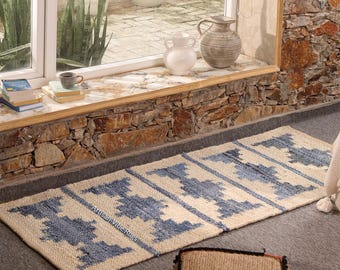 May include: A rectangular woven runner rug featuring a repeating blue geometric pattern on a natural beige background. The rug displays a textured surface and includes the text Artisan Vibe Rugs printed in a small font on the fabric surface.
