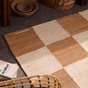May include: A rectangular area rug featuring a repeating checkerboard pattern of natural tan jute and off-white woven fibers. The rug displays the text DurrieIndiaCraftz printed in small white letters on one of the tan squares.