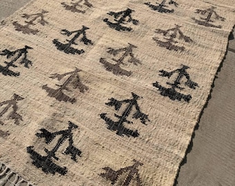 May include: A rectangular area rug featuring a cream-colored woven base with a repeating pattern of dark brown, abstract geometric symbols arranged in a grid. The rug is finished with short, knotted fringe along the top and bottom edges.