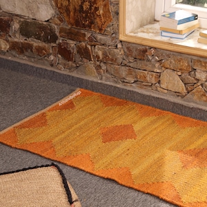 May include: An orange and yellow geometric patterned rug. The rug is rectangular and has a repeating diamond pattern. The rug is made of a woven material and is placed on a gray surface. The rug is near a stone wall and a window.