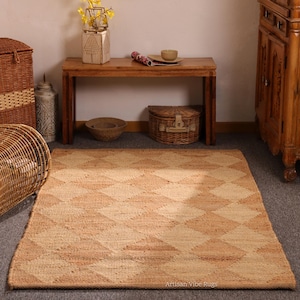 May include: A rectangular jute rug with a diamond pattern in shades of tan and beige. The rug is placed on a gray surface and is surrounded by wicker baskets and wooden furniture. The rug's texture is visible.