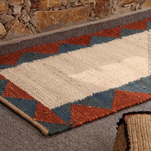 May include: A rectangular jute rug with a cream-colored center and a border of alternating blue and rust-colored triangles. The rug is placed on a gray surface near a stone wall. The text "ARTISANVIBERUGS" is visible.