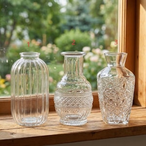 Könnte beinhalten: Drei klare Glasvasen in verschiedenen Designs stehen auf einer Holzfensterbank. Die Vasen umfassen eine gerippte, eine rautenförmige und eine sternförmige Vase. Der Hintergrund zeigt eine verschwommene Ansicht von Grün.