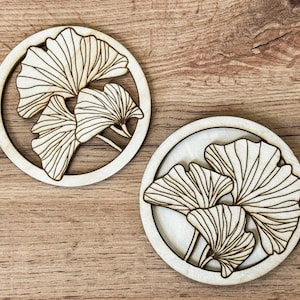 Pode incluir: Duas bases de madeira redondas com um design de folha de ginkgo cortada a laser. As folhas são castanho-claras com padrões de veias detalhados, dentro de uma moldura circular. As bases estão sobre uma superfície de madeira.