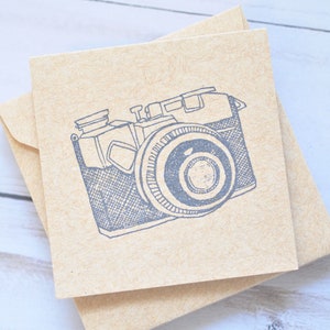 May include: A brown card with a blue illustration of a vintage camera. The camera has a lens with a circular shape and a textured body.