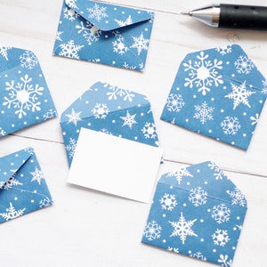 May include: Five small blue envelopes featuring a white snowflake pattern, arranged in a circular formation. One envelope in the center is open and contains a blank white rectangular card. A black pen is positioned in the upper right corner.