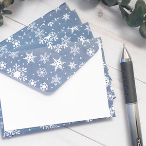 May include: A stack of blue envelopes featuring a white snowflake pattern, each with a triangular flap and a plain white rectangular card tucked inside. A black and silver mechanical pen is positioned to the right of the stationery set.