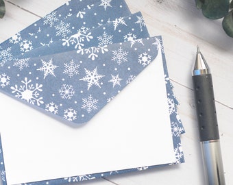 May include: A stack of blue envelopes featuring a white snowflake pattern, each with a triangular flap and a plain white rectangular card tucked inside. A black and silver mechanical pen is positioned to the right of the stationery set.