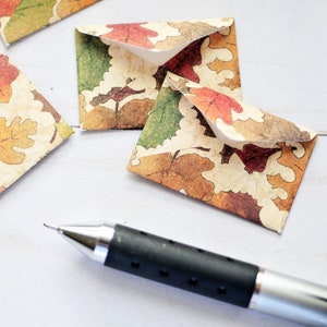 May include: Small, square paper envelopes with a fall leaf pattern in shades of red, orange, green, and brown. A silver and black pen is in the foreground. The envelopes are open and closed.