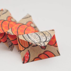 May include: A series of small, folded paper envelopes. The envelopes are decorated with a repeating pattern of orange pumpkins on a brown background. Some envelopes have a vintage newspaper print. The envelopes are arranged in a chain.