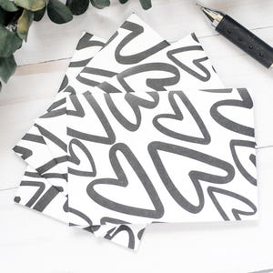 May include: A stack of white notecards with a repeating pattern of gray hearts. A silver and black pen rests on a white wooden surface, with a sprig of green leaves in the background.