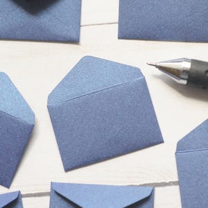 May include: A close-up of several small blue envelopes on a white surface. A black pen is lying on the surface.