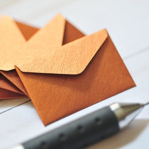 May include: A stack of small, folded, copper-colored envelopes on a white surface. A black pen is in the foreground.