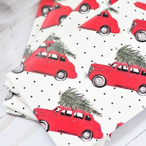May include: A stack of square paper napkins with a repeating pattern of red vintage cars carrying Christmas trees on a white background with black polka dots. The napkins are folded and stacked on a light wood surface.