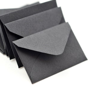 May include: A stack of black envelopes, with the top envelope in focus. The envelopes are made of a textured paper and have a simple, classic design.