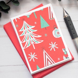 May include: A stack of Christmas cards with a red background and a pattern of white and teal Christmas trees, ornaments, and snowflakes. The cards are on top of a red envelope. A black and silver pen rests on the right.